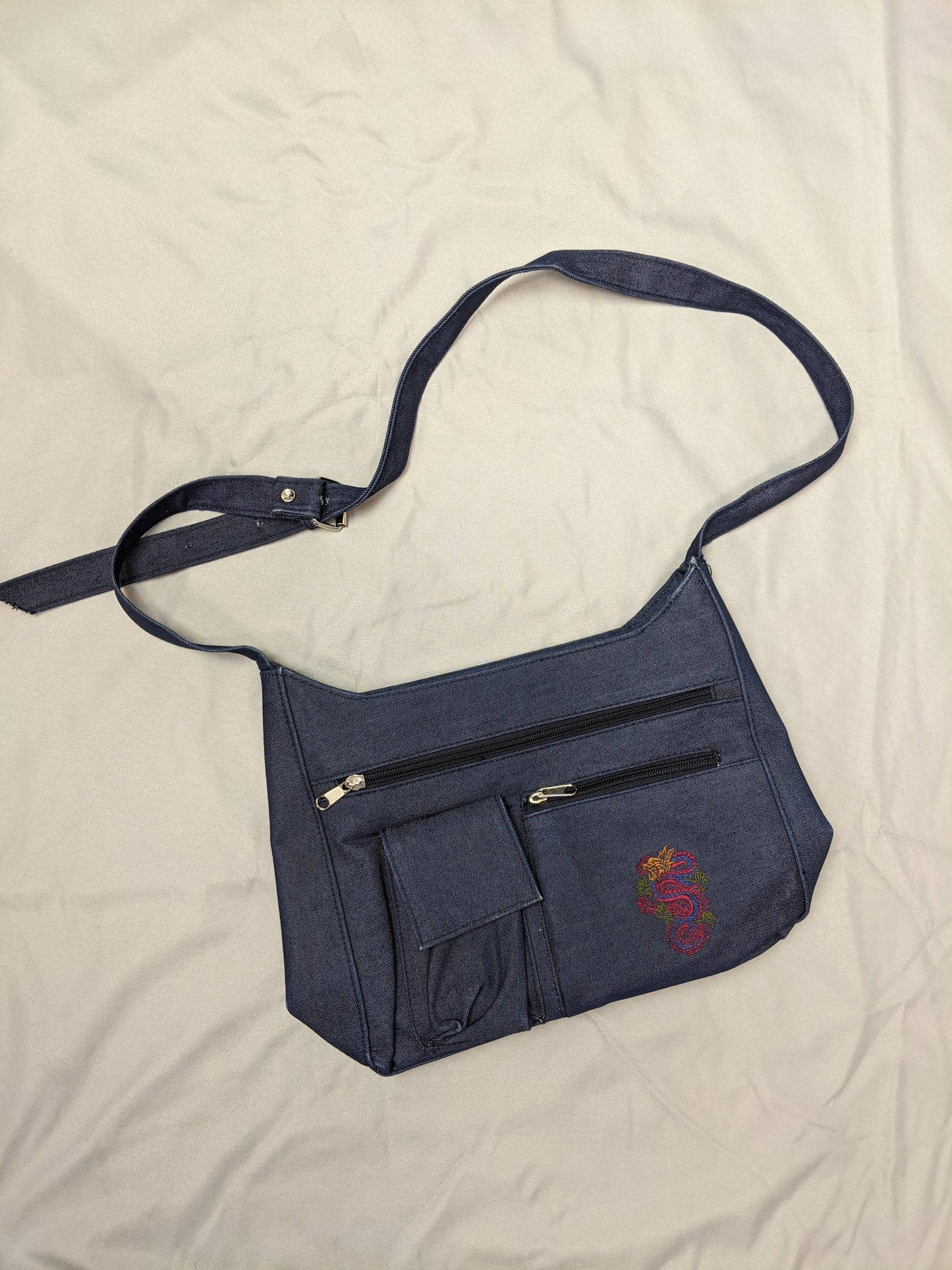 Blue denim shoulder bag with dragon embroidery and pockets on beige background.