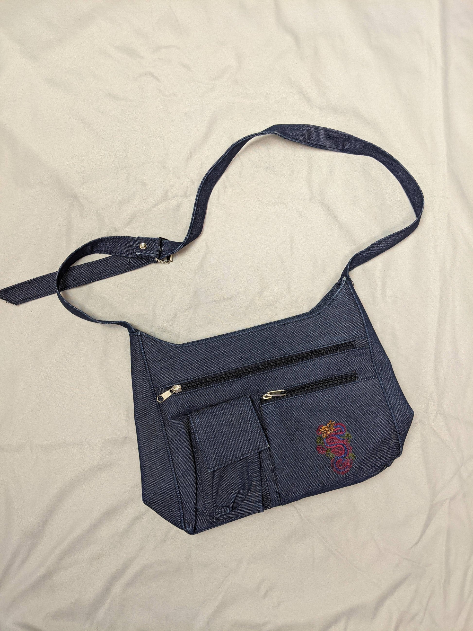 Blue denim shoulder bag with dragon embroidery and pockets on beige background.