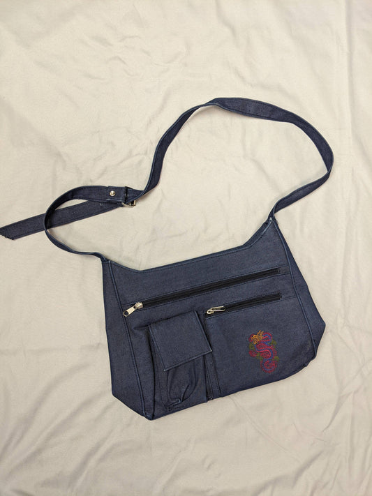 Blue denim shoulder bag with dragon embroidery and pockets on beige background.
