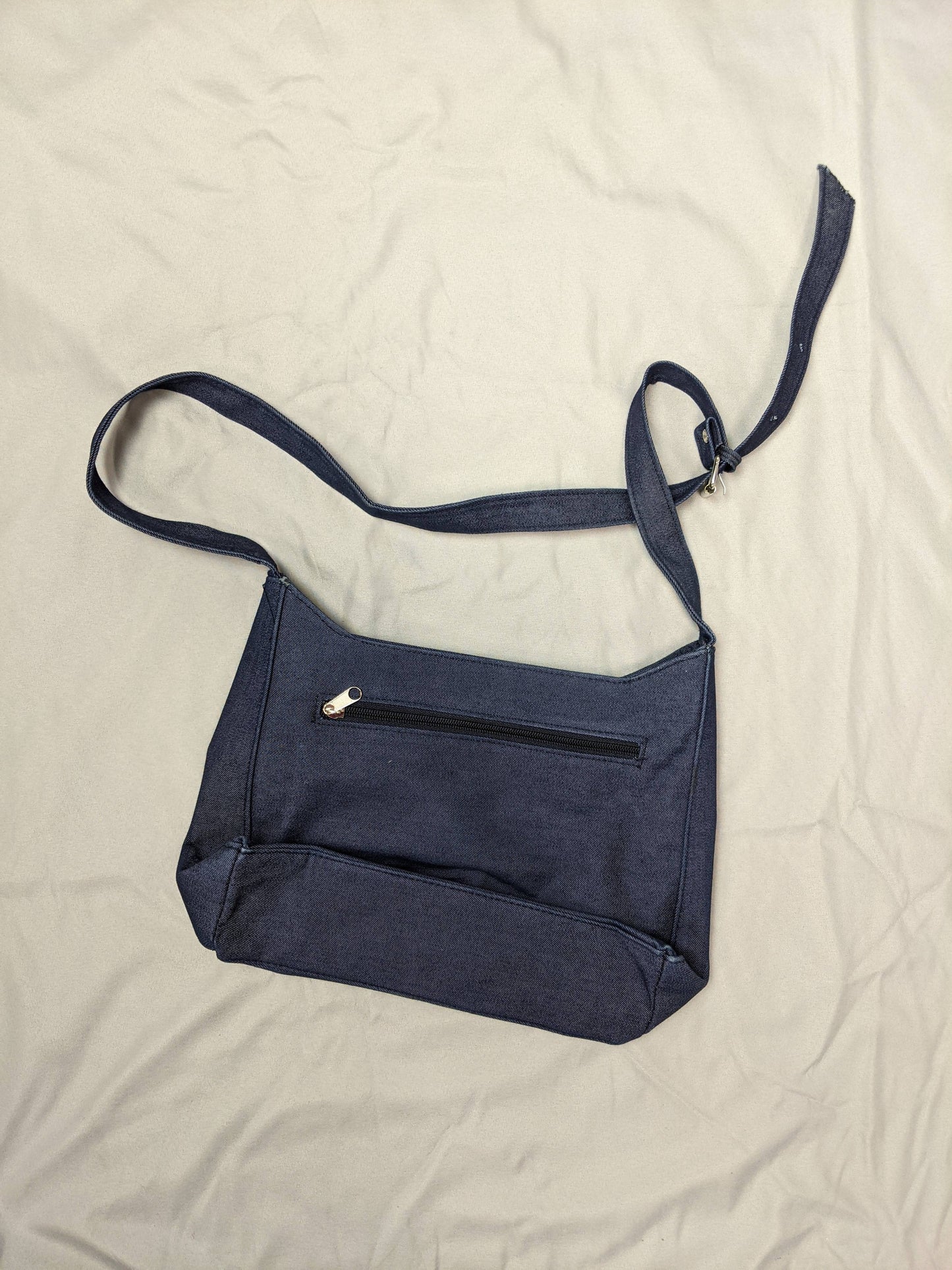 Back view of blue denim shoulder bag with back zipper pocket on beige background.