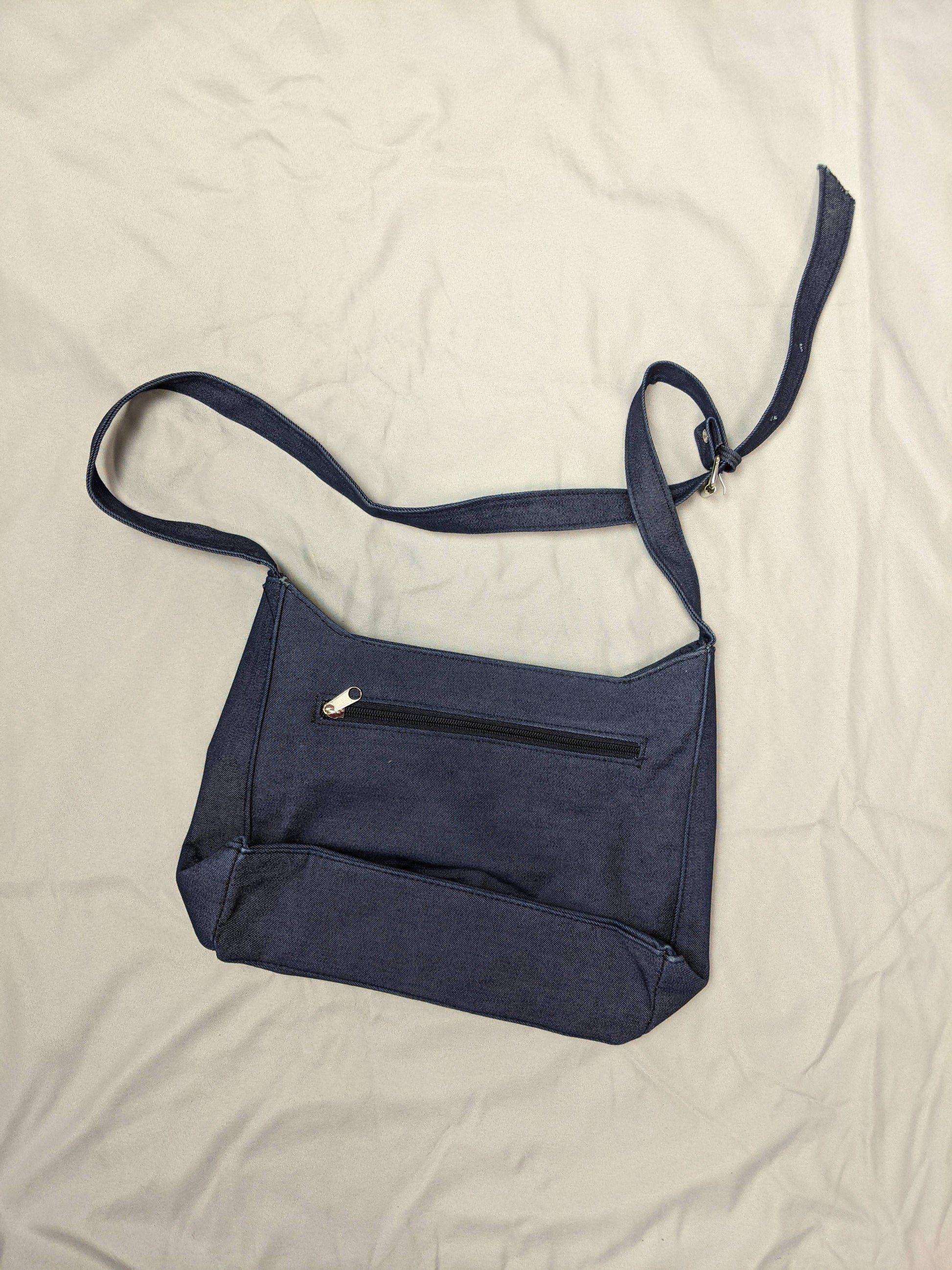 Back view of blue denim shoulder bag with back zipper pocket on beige background.