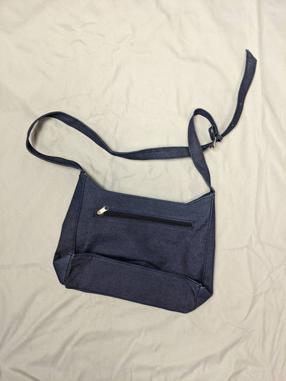 Back view of blue denim shoulder bag with back zipper pocket on beige background.